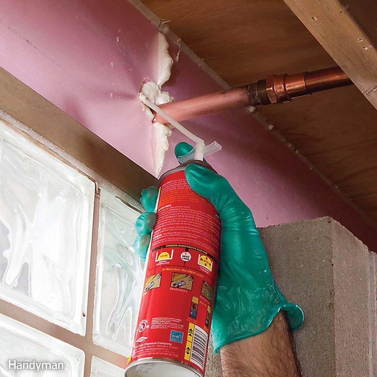 A gloved hand holds a spray can, applying foam sealant to a gap around a copper pipe. The setting features a wooden beam and glass brick wall.