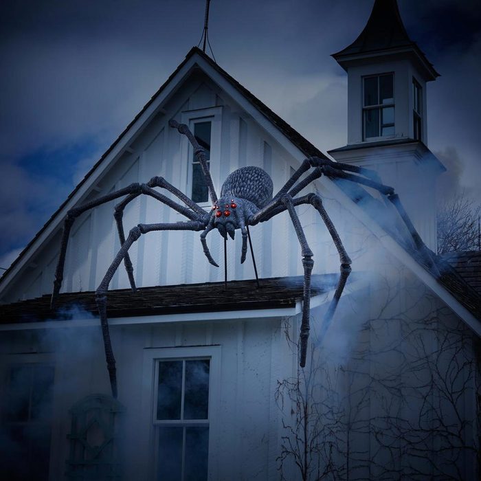 A large, decorative spider with glowing red eyes descends from the roof of a white house, surrounded by mist and a dark, cloudy sky.