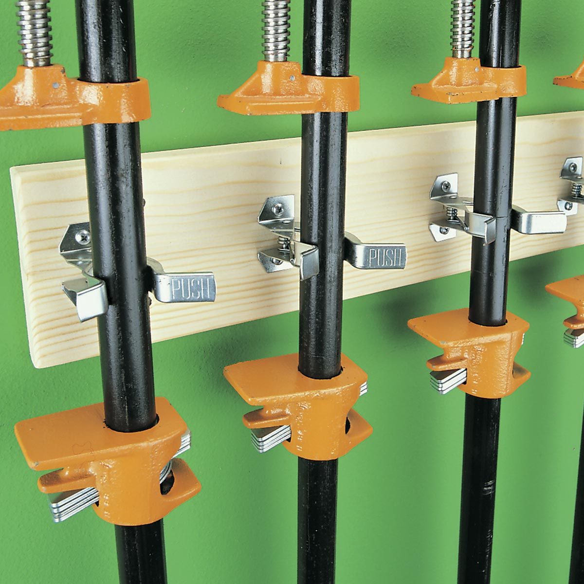Clamps with orange pads are mounted on black pipes attached to a wooden board, secured against a green wall, ready for use.