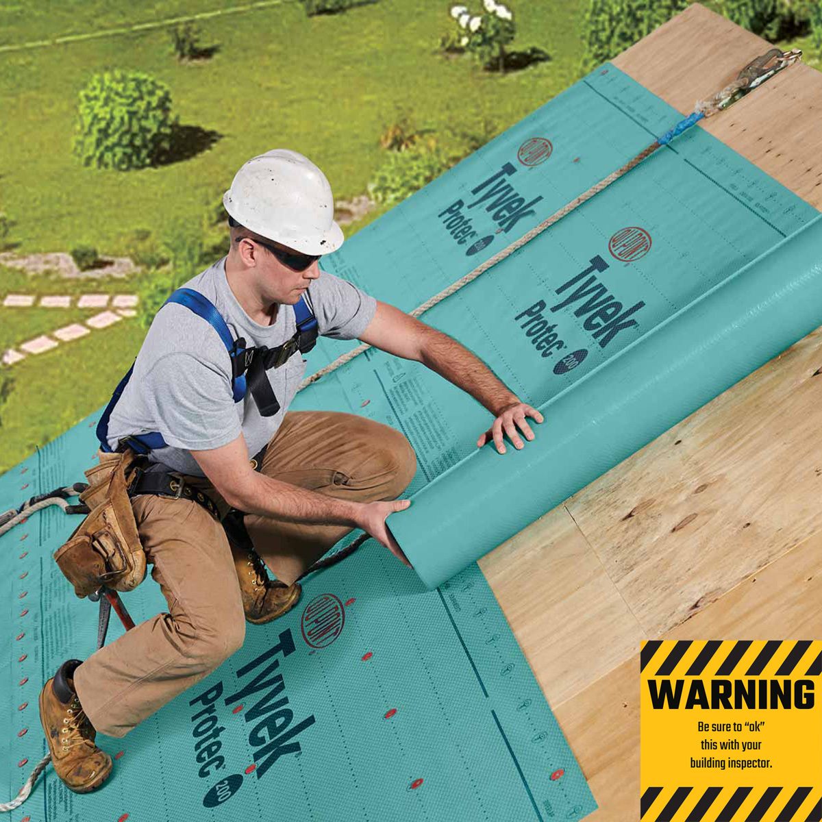 A worker applies a large blue Tyvek protective material on a roof. The scene is outdoors, surrounded by green grass and trees, indicating a construction site.