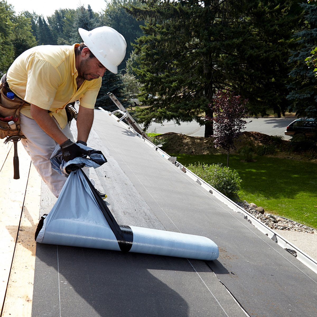 A man in a hard hat rolls out roofing material while standing on a sloped roof, surrounded by trees and a well-maintained lawn.