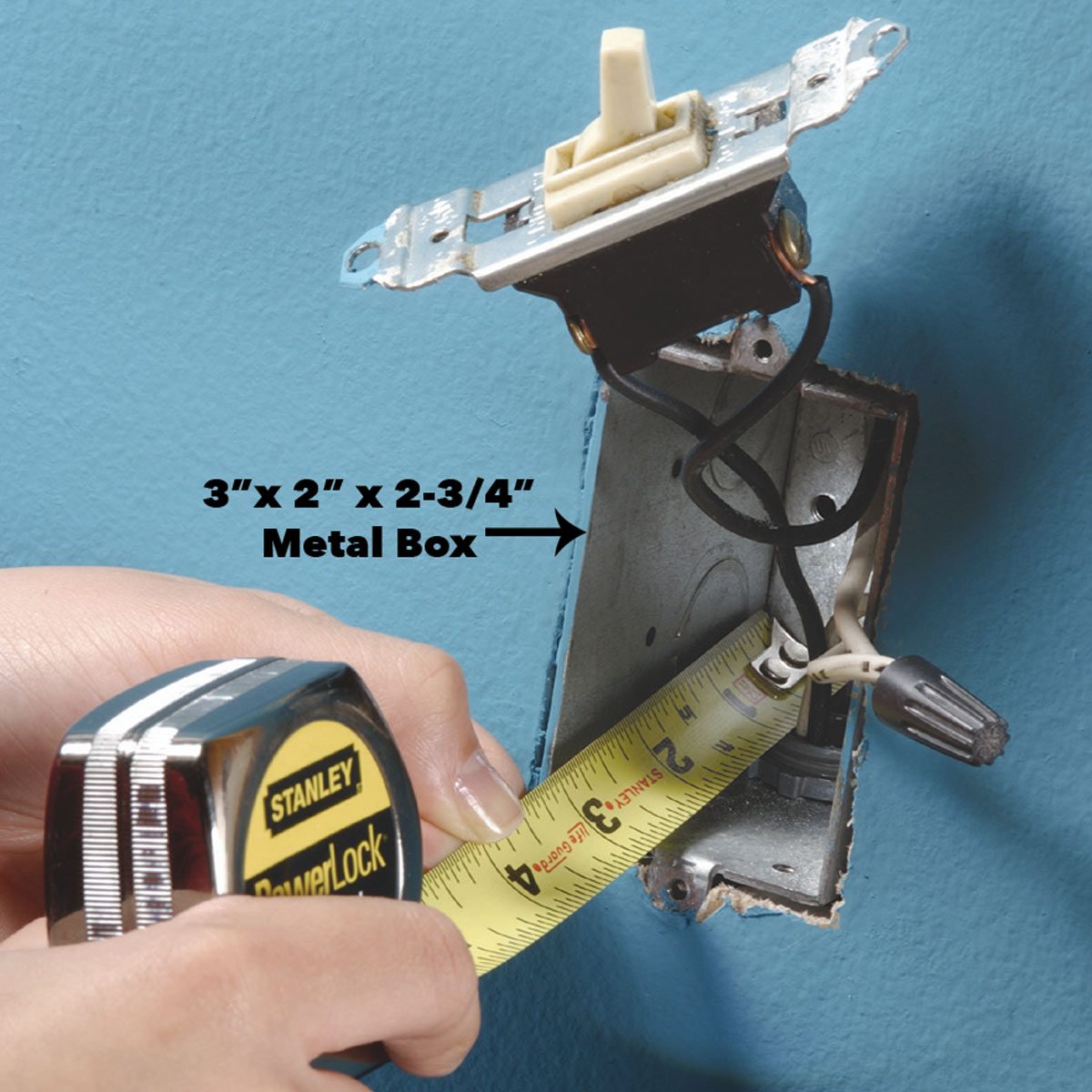 A person measures a metal electrical box, revealing electrical wires inside, against a blue wall. A measuring tape indicates dimensions of 3” x 2” x 2-3/4”.