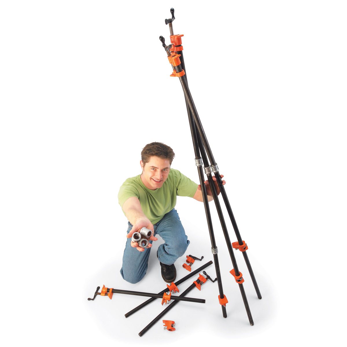 A man holds metal connectors while assembling a tripod structure using black rods and orange clamps on a white background. He appears engaged and smiling.