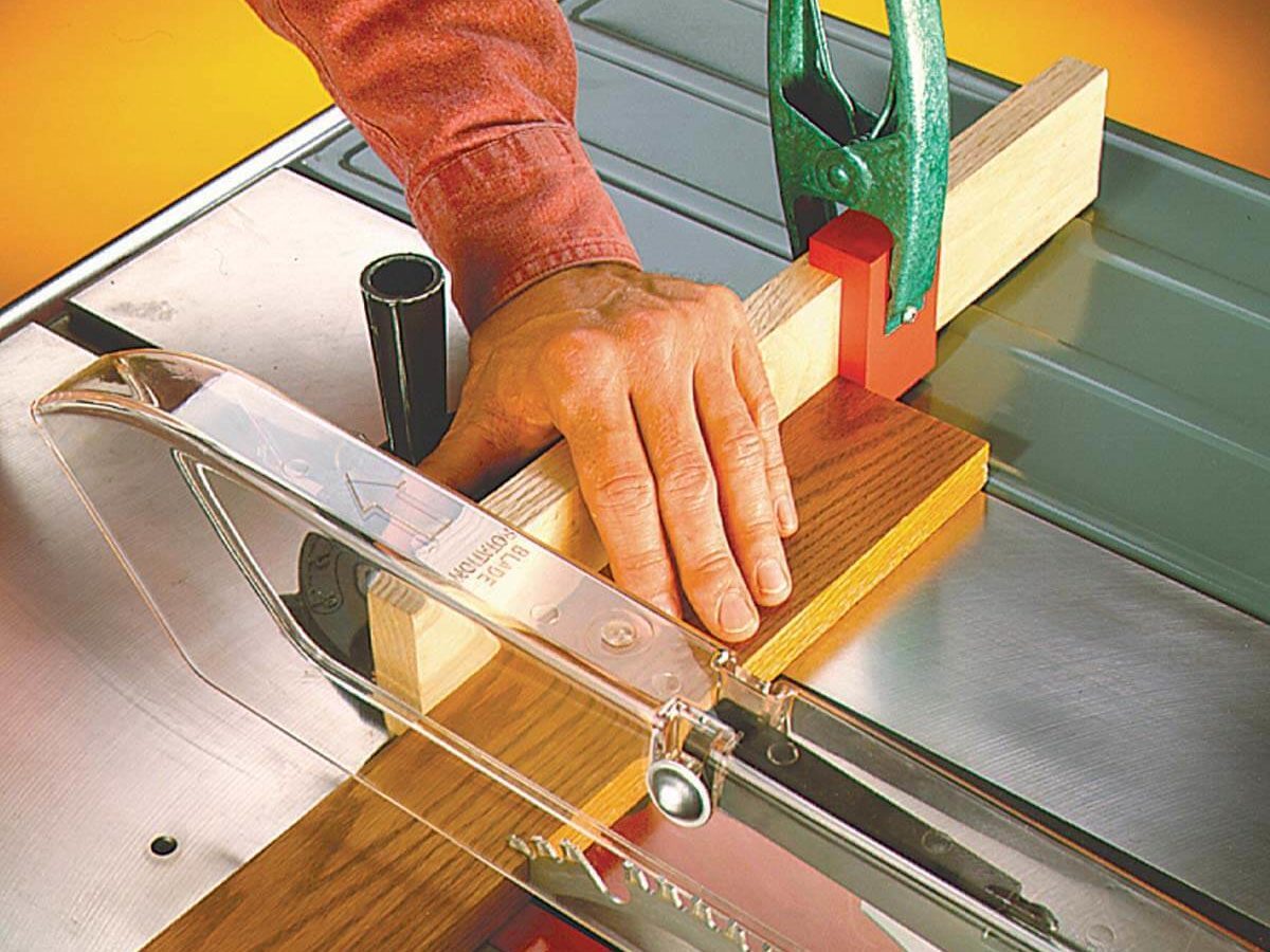 A hand positions a wooden block against a blade on a table saw, secured with a clamp, focused on precise cutting in a workshop environment.