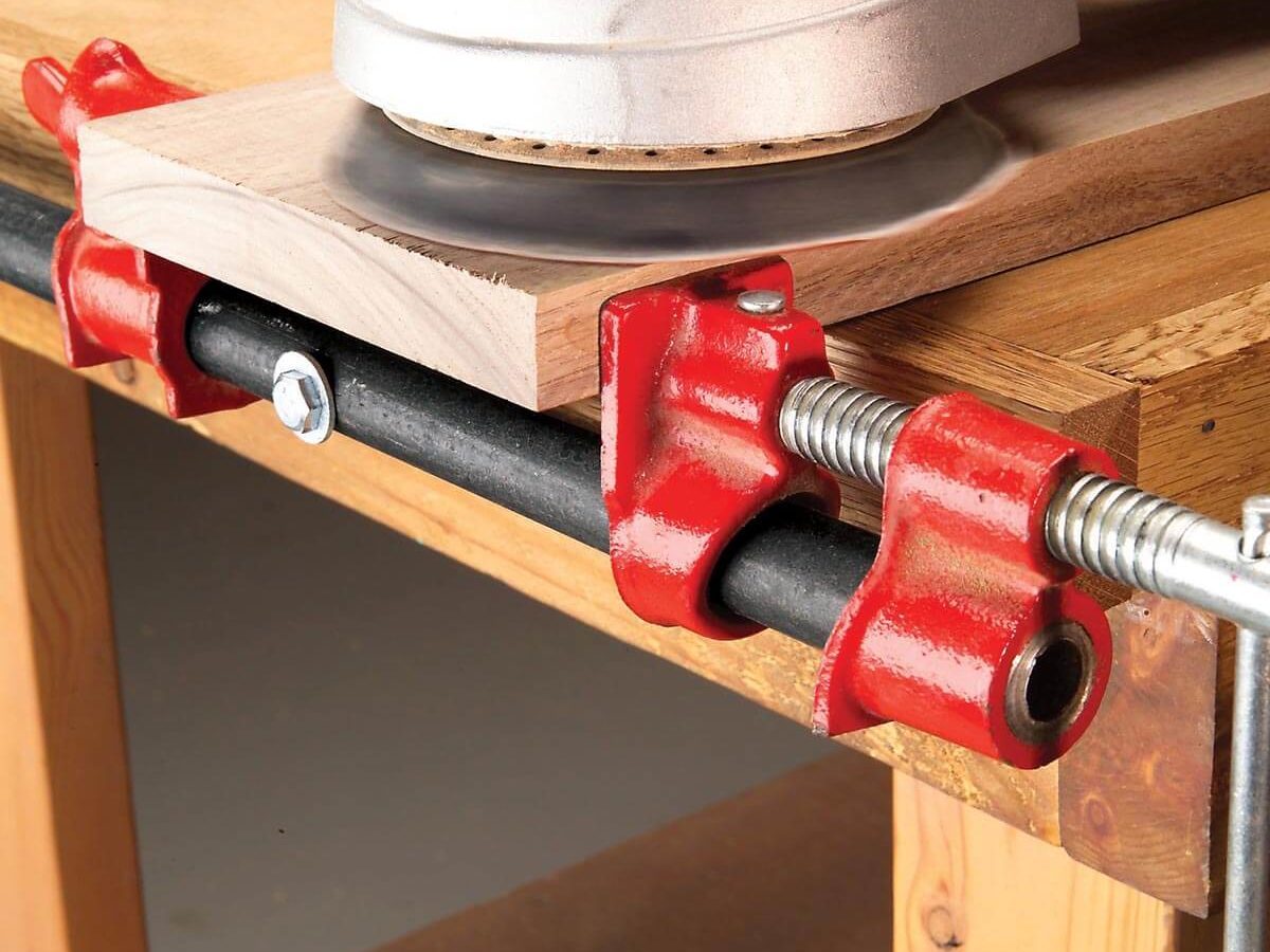 A power sander is used on a piece of wood, secured by red clamps on a workbench, highlighting a woodworking task in progress.