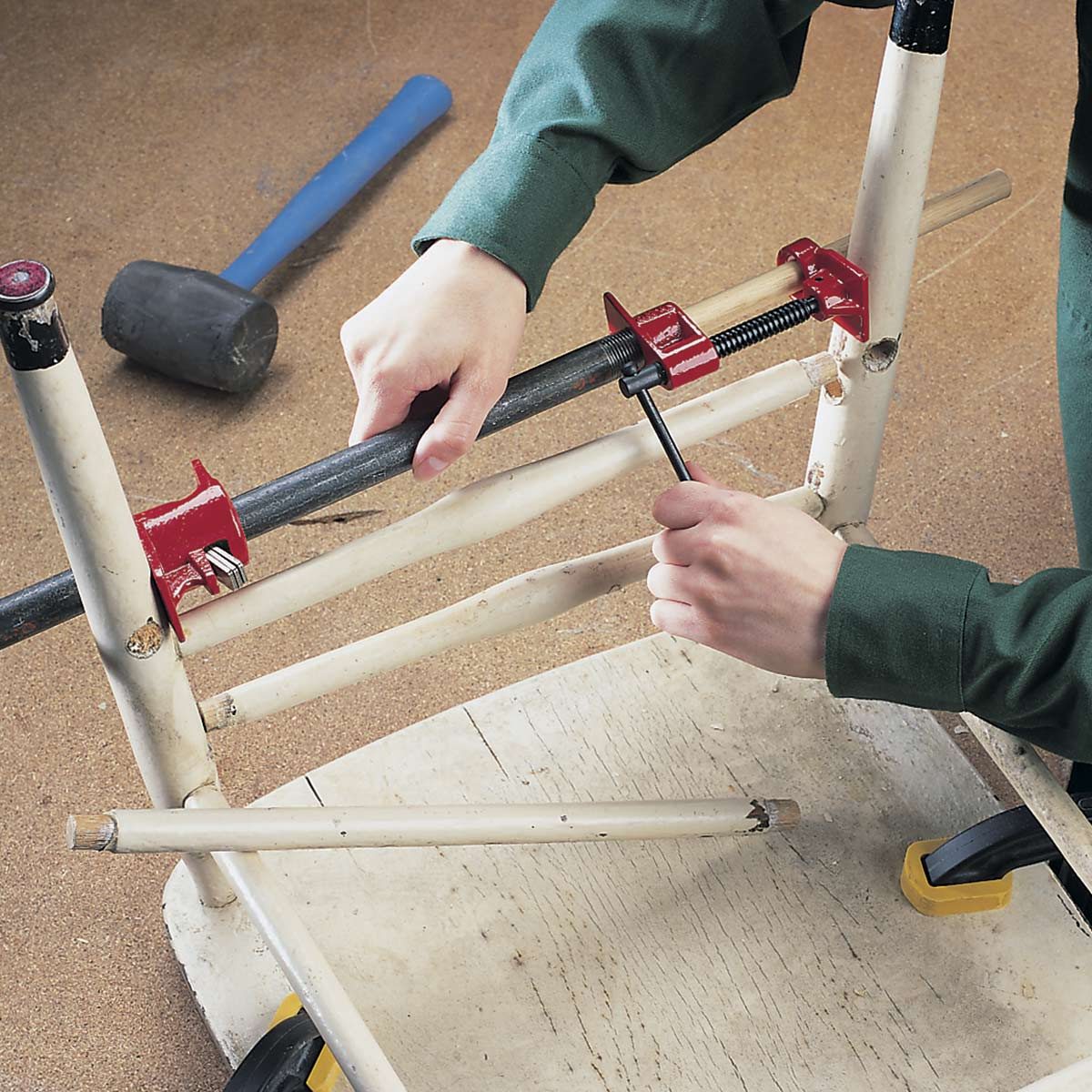 A person is tightening a clamp on a metal rod, securing it to a wooden chair frame on a work surface, with a rubber mallet nearby.