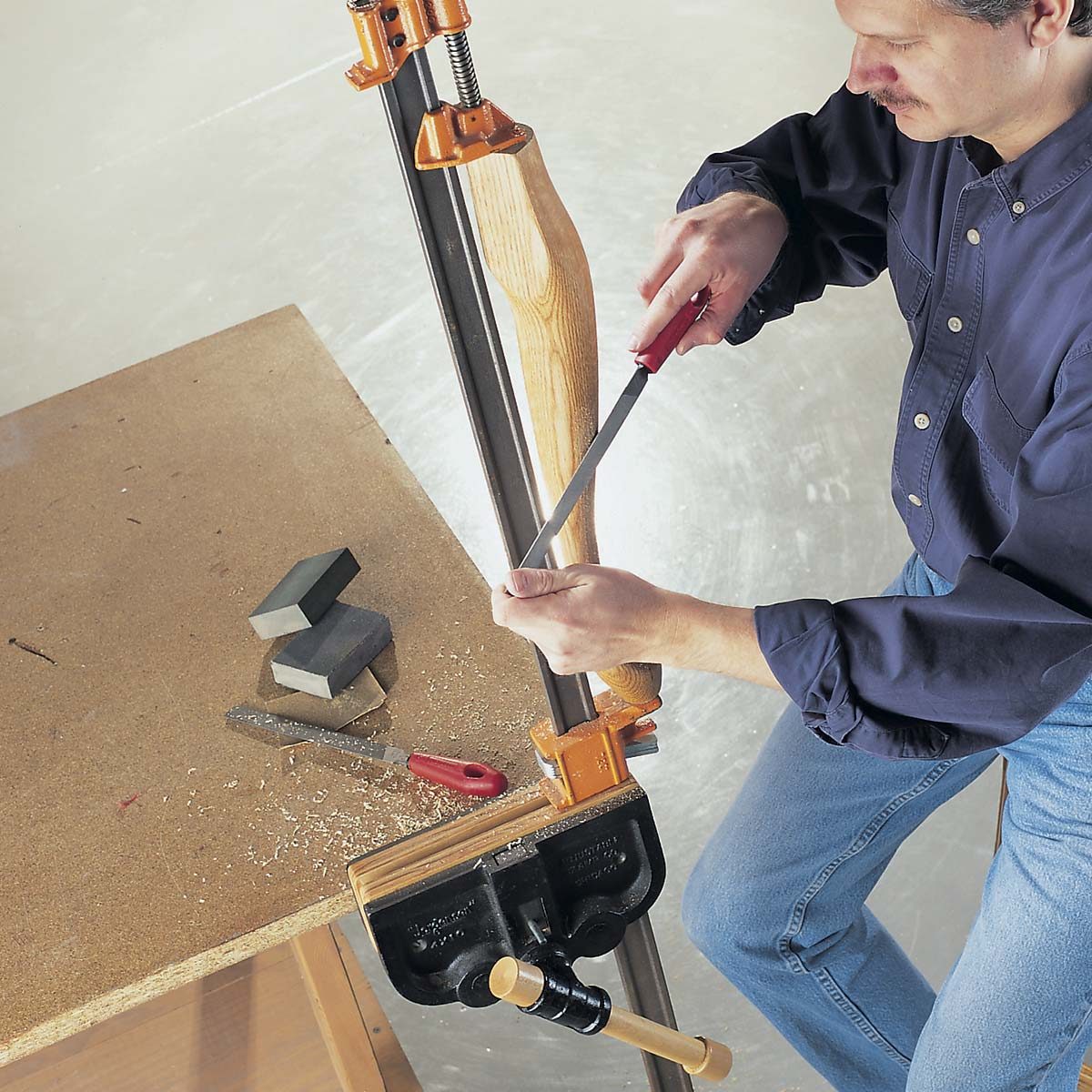 A person uses a file on a wooden object secured in a clamp on a workbench, surrounded by woodworking tools and shavings.