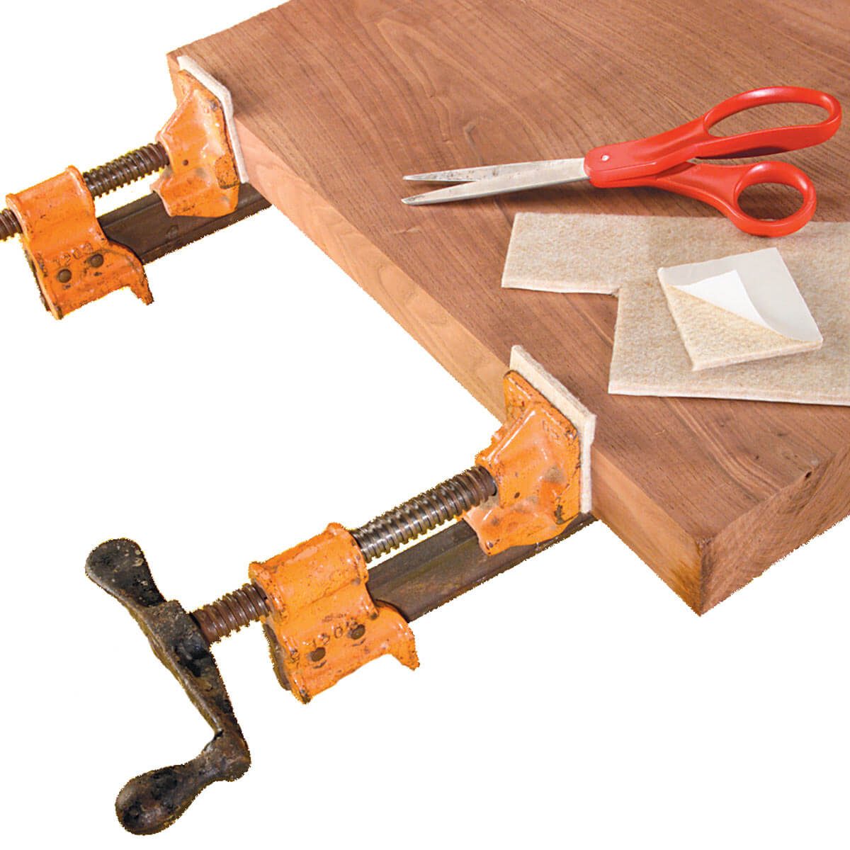 Wooden board is clamped by two orange clamps. Scissors and pieces of material are placed nearby on the surface, suggesting preparation for a craft project.