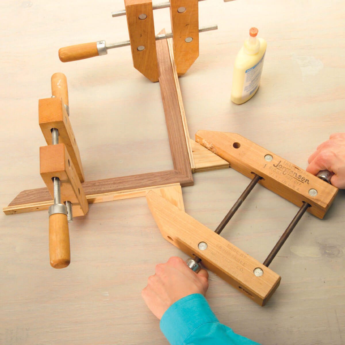 A person is tightening two clamps on a wooden frame, applying adhesive, with other tools visible on a light-colored work surface.
