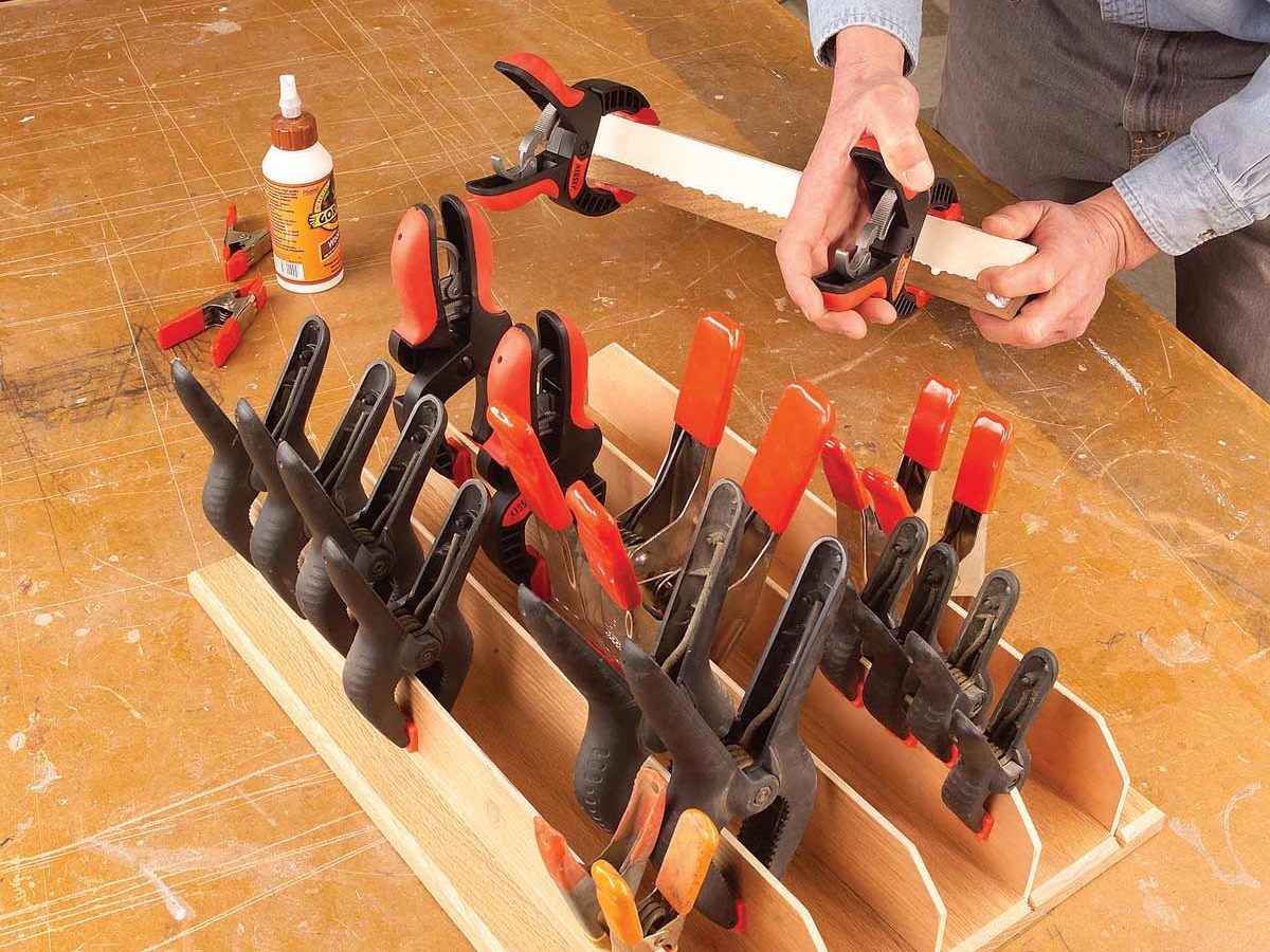 A person is applying pressure using a clamp on a wooden piece, while various clamps are organized in a holder on a workbench. A glue bottle sits nearby.