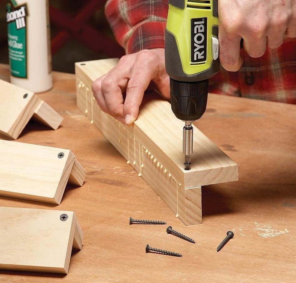 A person uses a power drill to secure a wooden piece with screws while surrounded by additional wood blocks and scattered screws on a workbench.