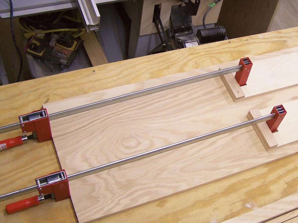 Clamps secure two pieces of wood on a workbench, providing stability for woodworking tasks in a workshop environment, surrounded by tools and materials.