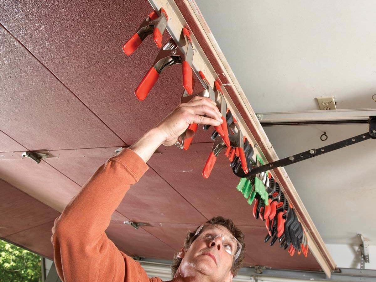 A man with glasses reaches up to adjust various clamps mounted on a wall in a garage, surrounded by tools and natural light.