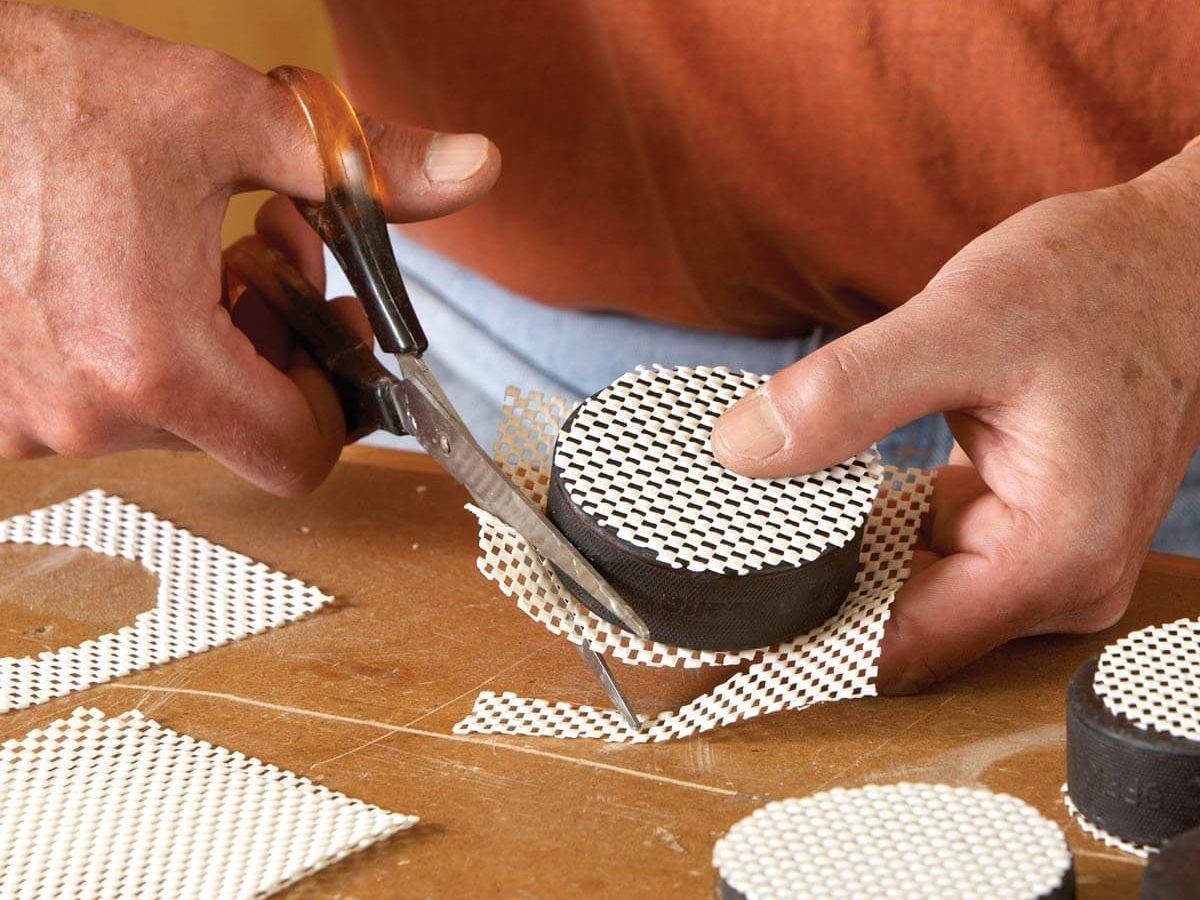 Hands are cutting a mesh material around a circular object on a wooden surface, with additional circular objects nearby, in a workshop setting.