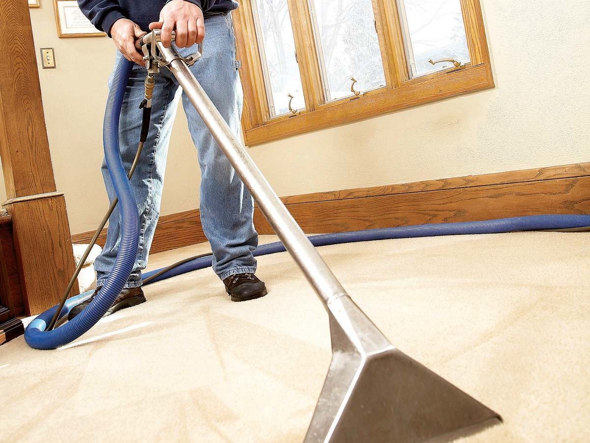 A person uses a carpet cleaning machine on light-colored carpet in a room with wooden accents and large windows, ensuring the surface is clean.