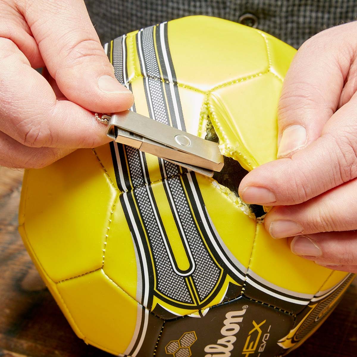 A yellow soccer ball is being repaired with a silver tool, as a person's hand holds the ball steady against a textured wooden surface.