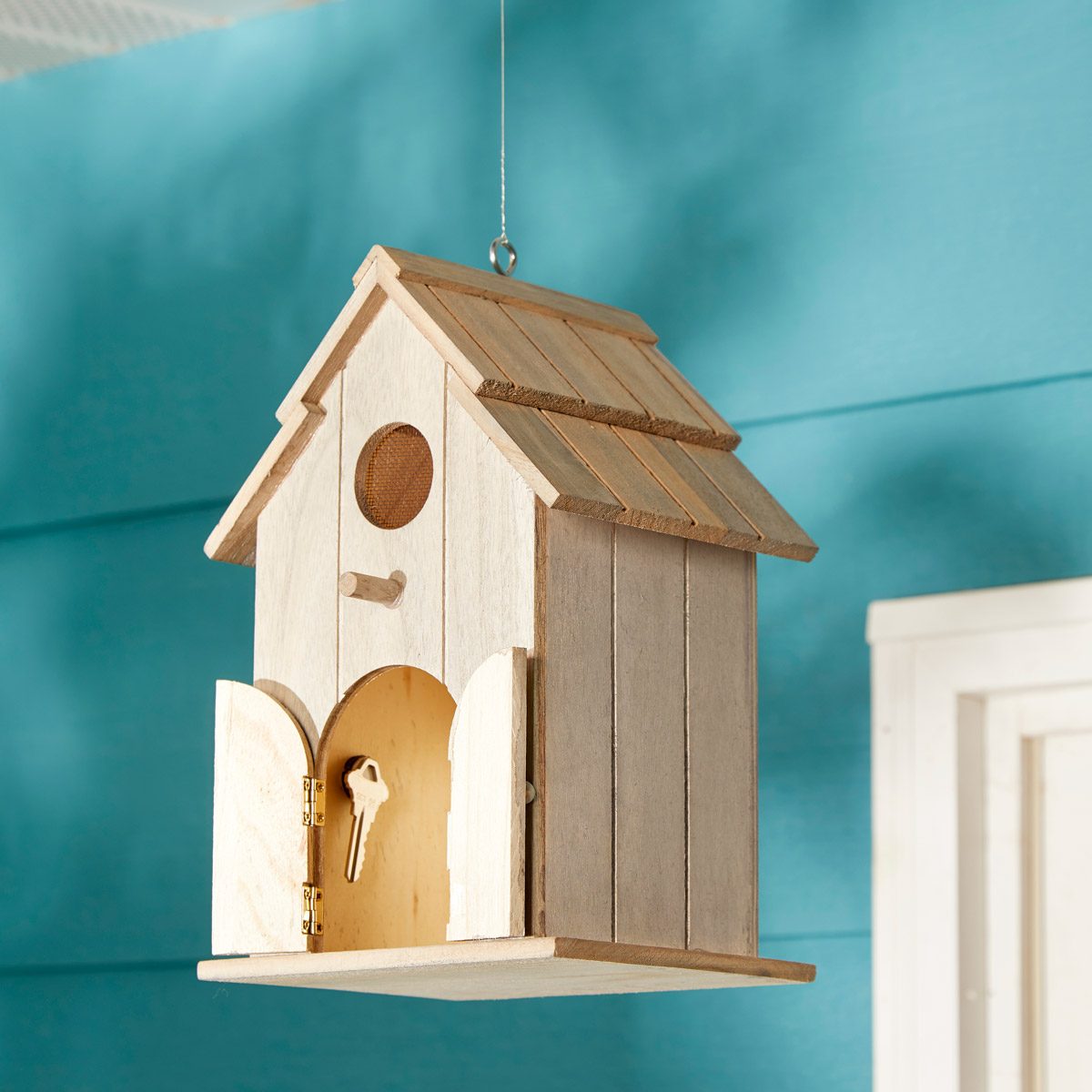 A wooden birdhouse hangs from a ceiling, featuring open doors and a key inside, against a bright blue wall.