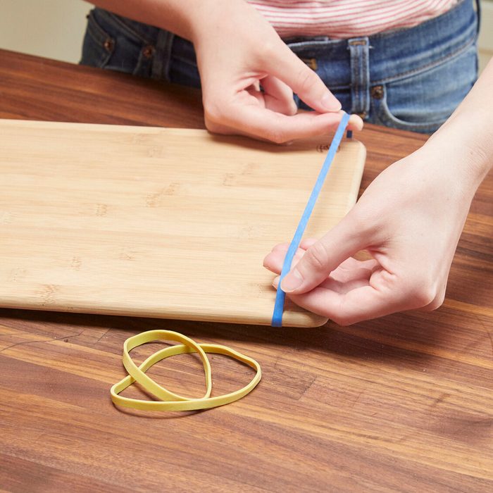 A person stretches a blue ribbon across a bamboo cutting board, while yellow rubber bands lie on the wooden surface nearby.