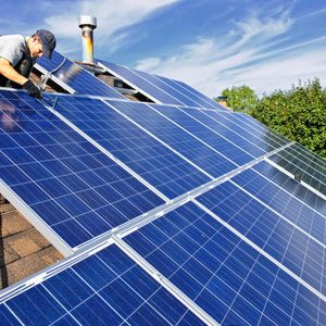 A technician installs solar panels on a sloped rooftop, surrounded by trees under a clear sky with scattered clouds.