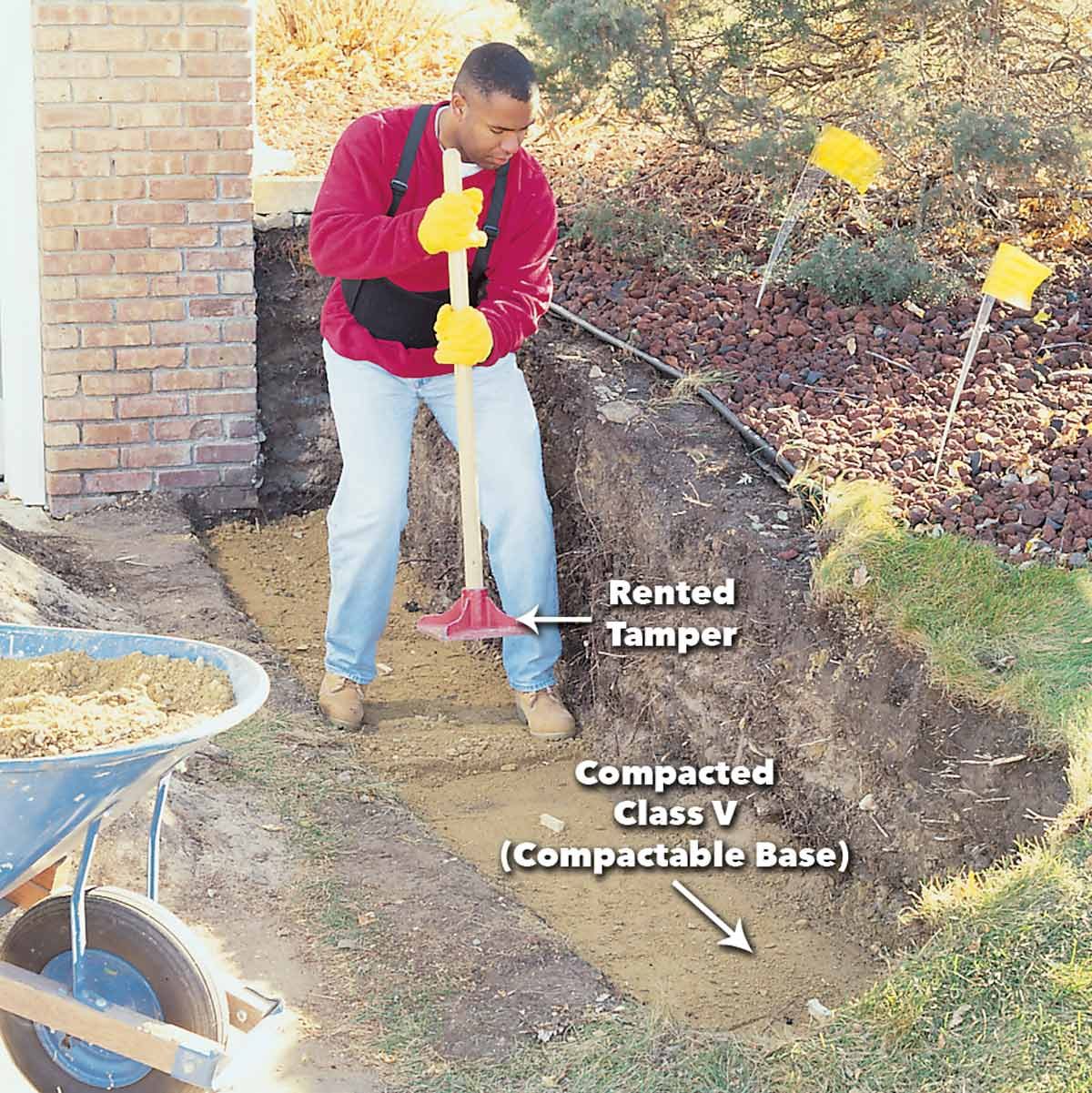 A man uses a tamper to compact soil in a trench beside a brick wall, with a wheelbarrow nearby and landscaping in the background.