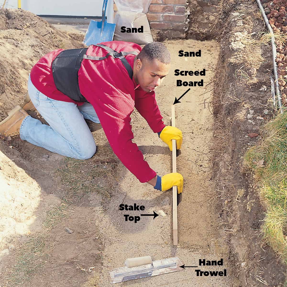 A person kneels in a trench, leveling sand with a screed board amid construction debris in a yard, wearing gloves and a red shirt.