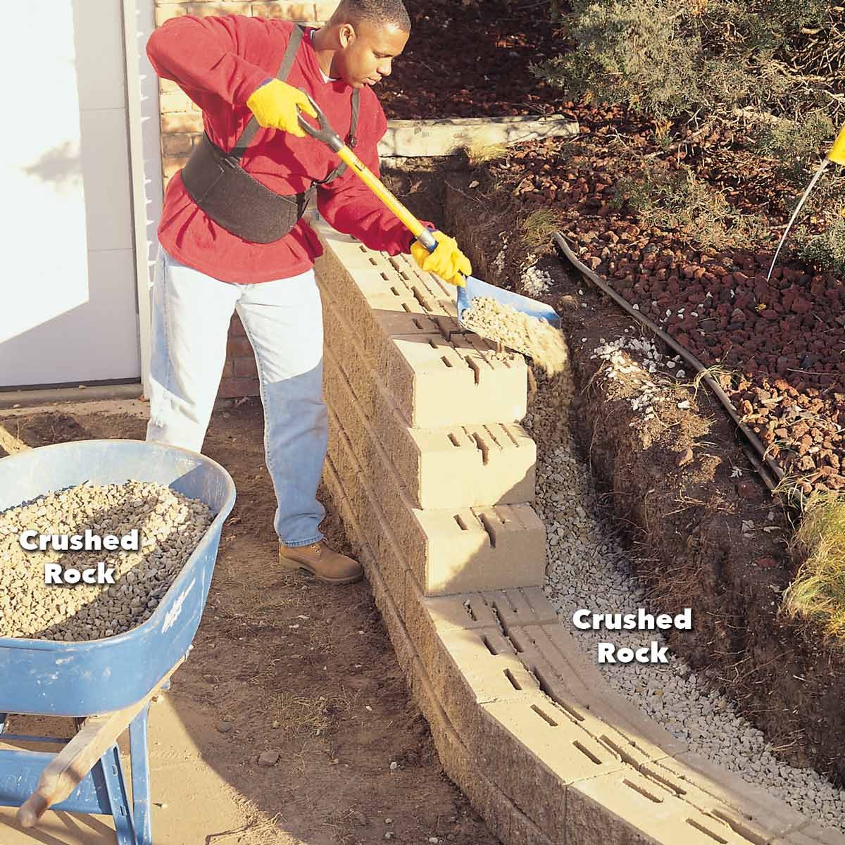 A person in a red shirt and gloves shovels crushed rock from a wheelbarrow onto a stone wall in a garden area.
