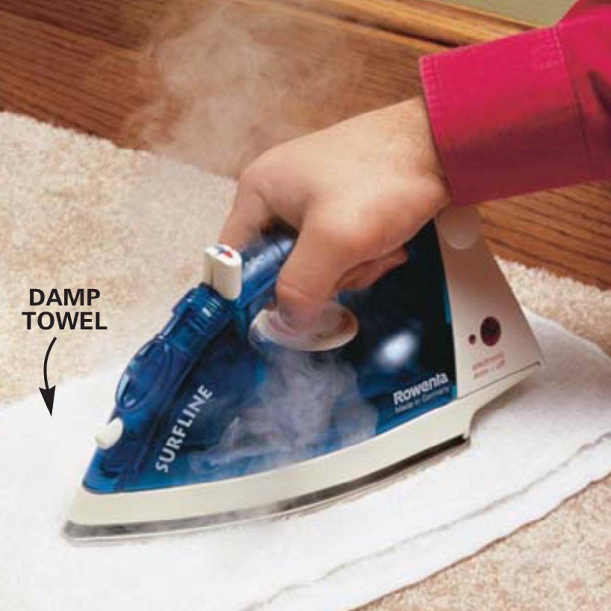 A hand holds a steaming iron over a damp towel placed on a carpet, actively pressing down to smooth the towel.