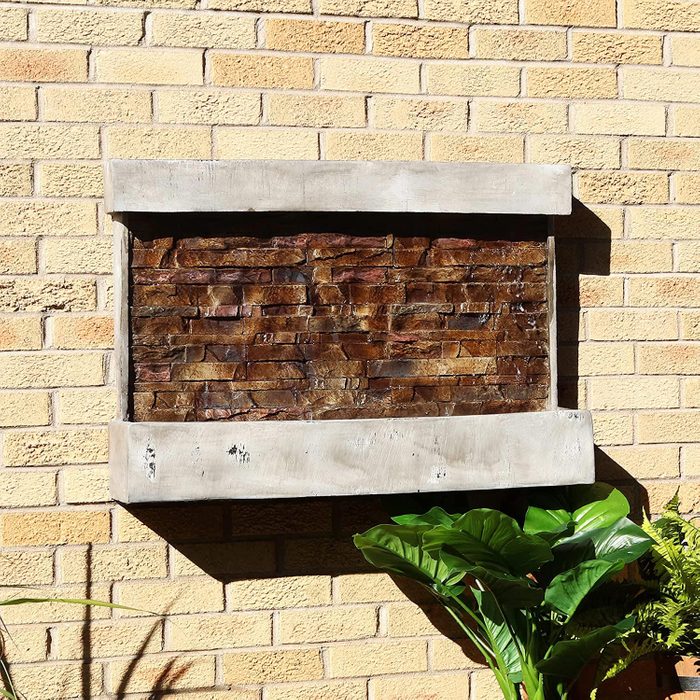 A rectangular wall-mounted fountain made of stacked stones trickles water, framed by a concrete border, against a yellow brick wall with potted greenery nearby.