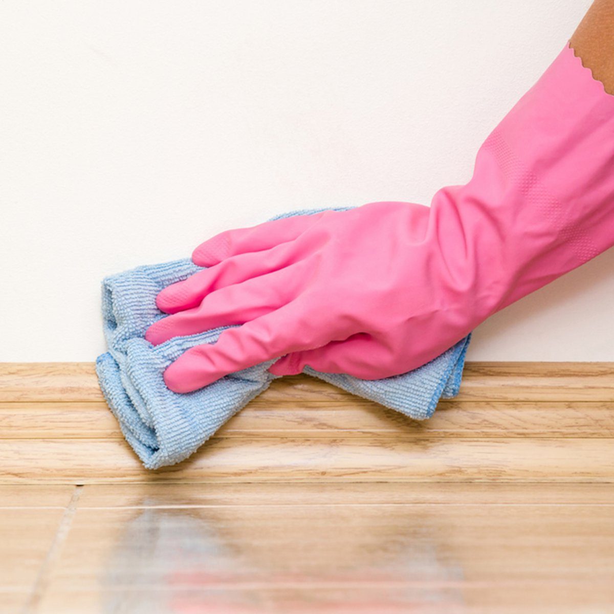A gloved hand wipes a blue cloth along a wooden baseboard in a clean, interior space. The wall behind is plain and light-colored.