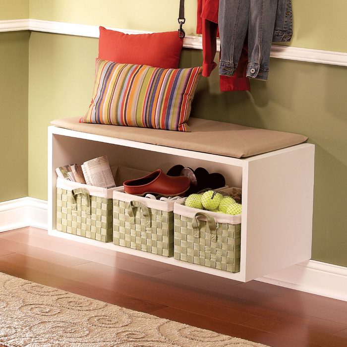 A white bench with a cushion holds a striped pillow, while beneath it, woven baskets contain shoes and various items, set against a green wall.