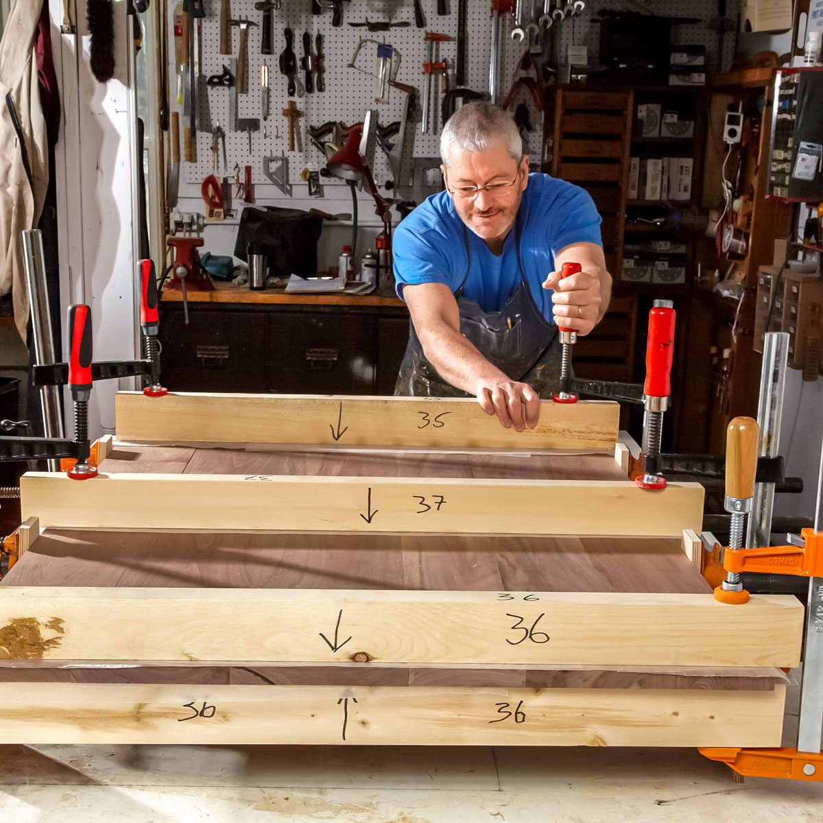A man tightens clamps on wooden boards laid out on a workbench in a well-equipped workshop, surrounded by tools and materials.