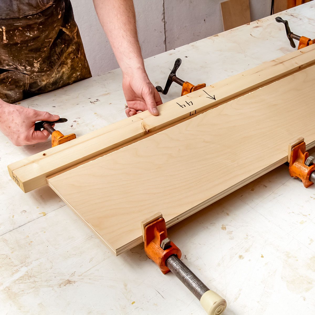 A person is positioning a wooden board against a larger piece, securing them with clamps on a workshop table. The environment is cluttered with woodwork materials.