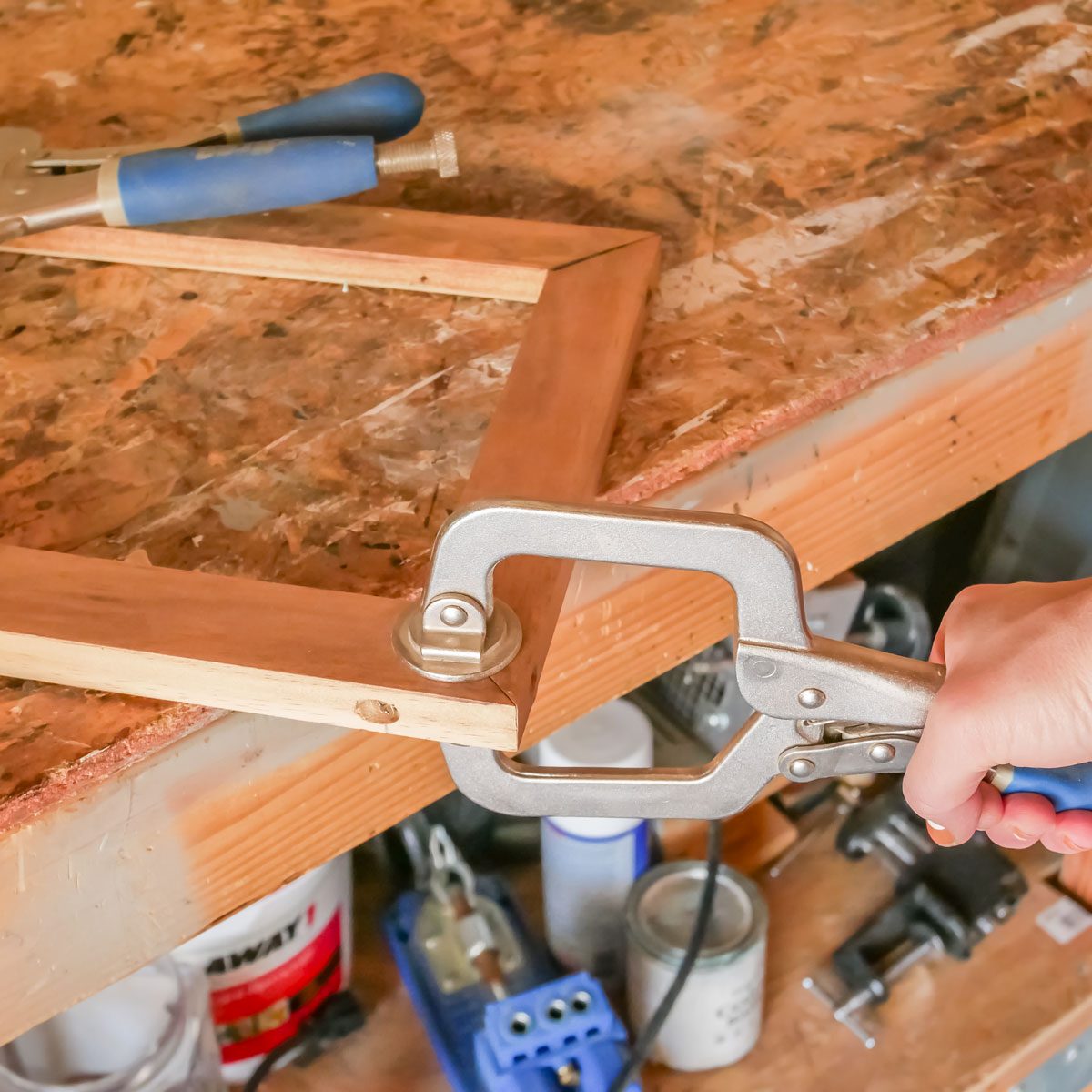 A hand grips a clamp tightening a wooden frame on a workbench, surrounded by various tools and supplies, indicating a woodworking task in progress.