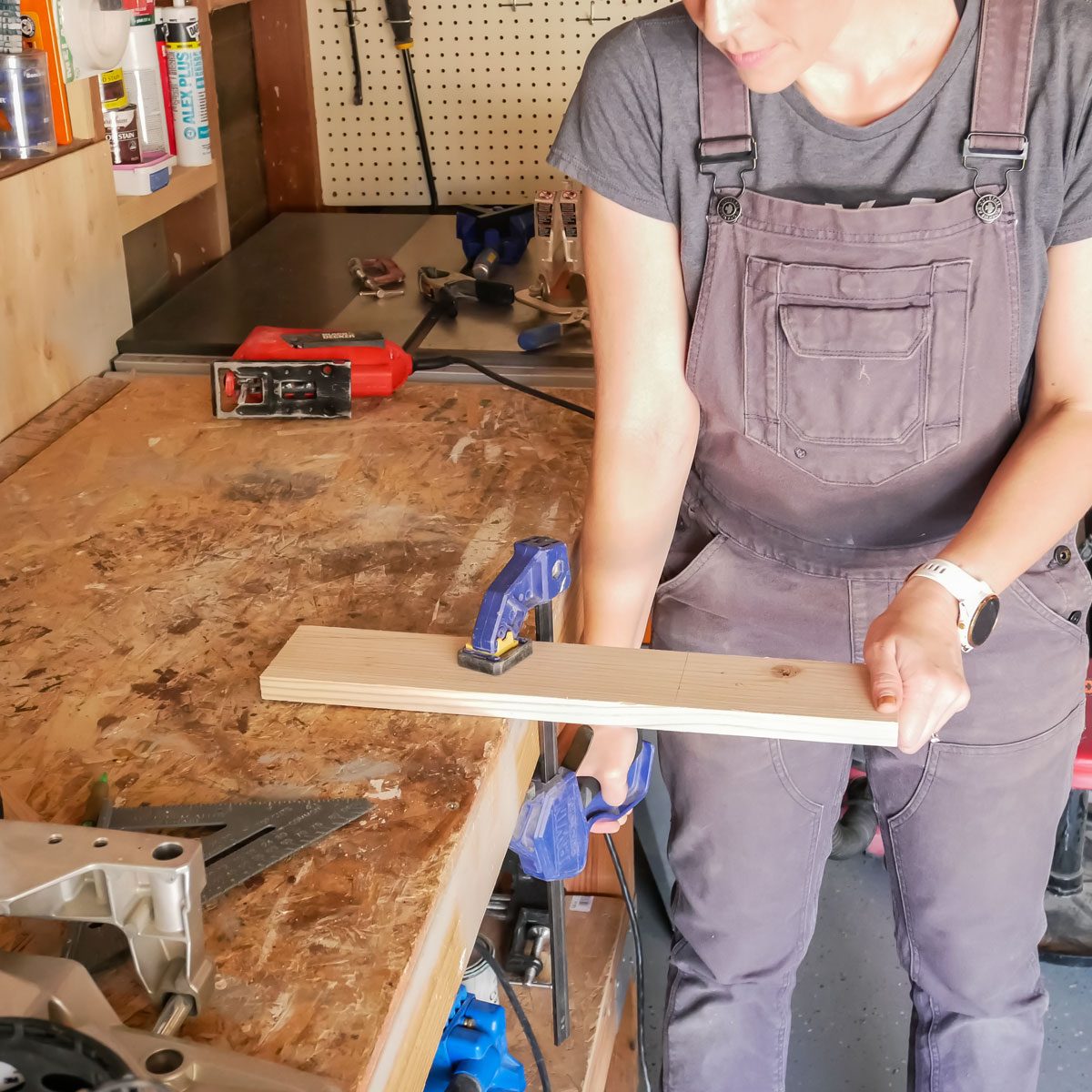 A person holds a wooden plank secured with a clamp, preparing to work in a well-equipped workshop with tools and materials around.