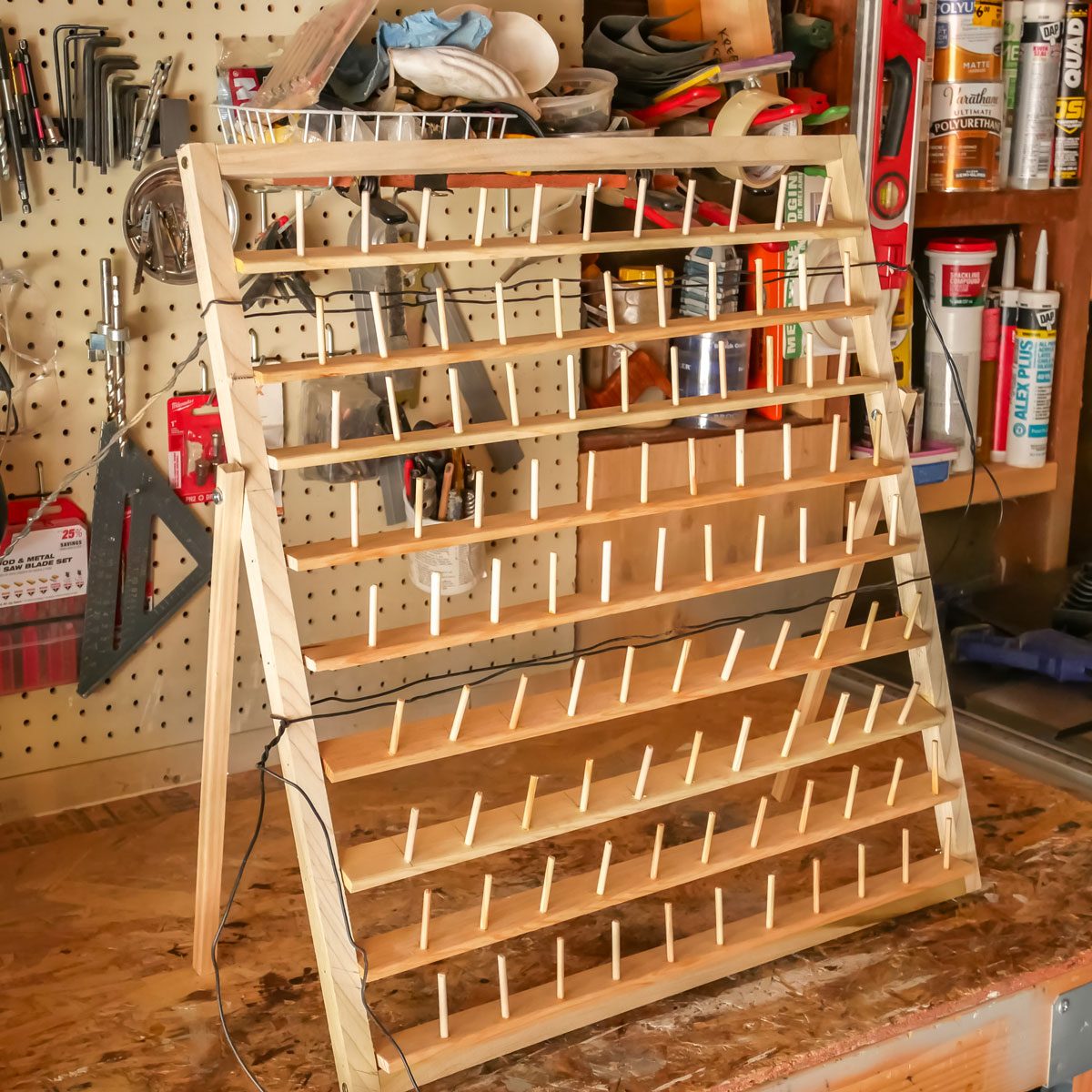 A wooden rack with multiple shelves stands upright, designed for organizing items, set against a backdrop of tools and supplies on a workshop wall.