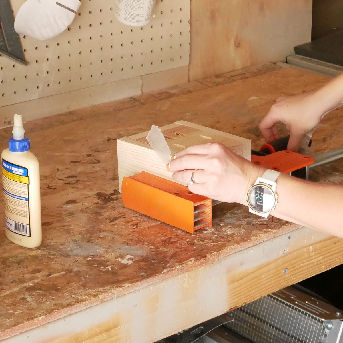 A person holds a piece of clear tape while working on a wooden block, surrounded by tools and a bottle of glue on a workbench.