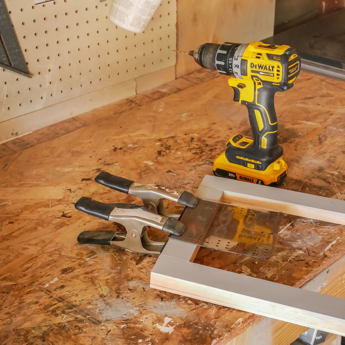 A power drill sits next to a clamp holding a metal piece against an unfinished wooden frame on an unfinished workbench in a workshop environment.