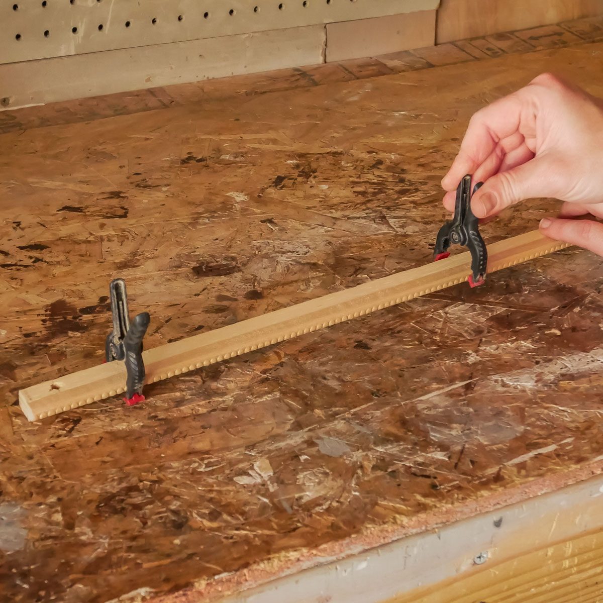 A hand adjusts a clamp on a wooden strip, securing it to a work surface made of oriented strand board, in a workshop setting.