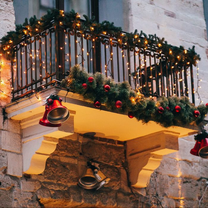 Stylish christmas decorations, red jingle bells, lights, fir branches with ornaments on balcony in european city street. Festive decor and illumination in city center, winter holidays