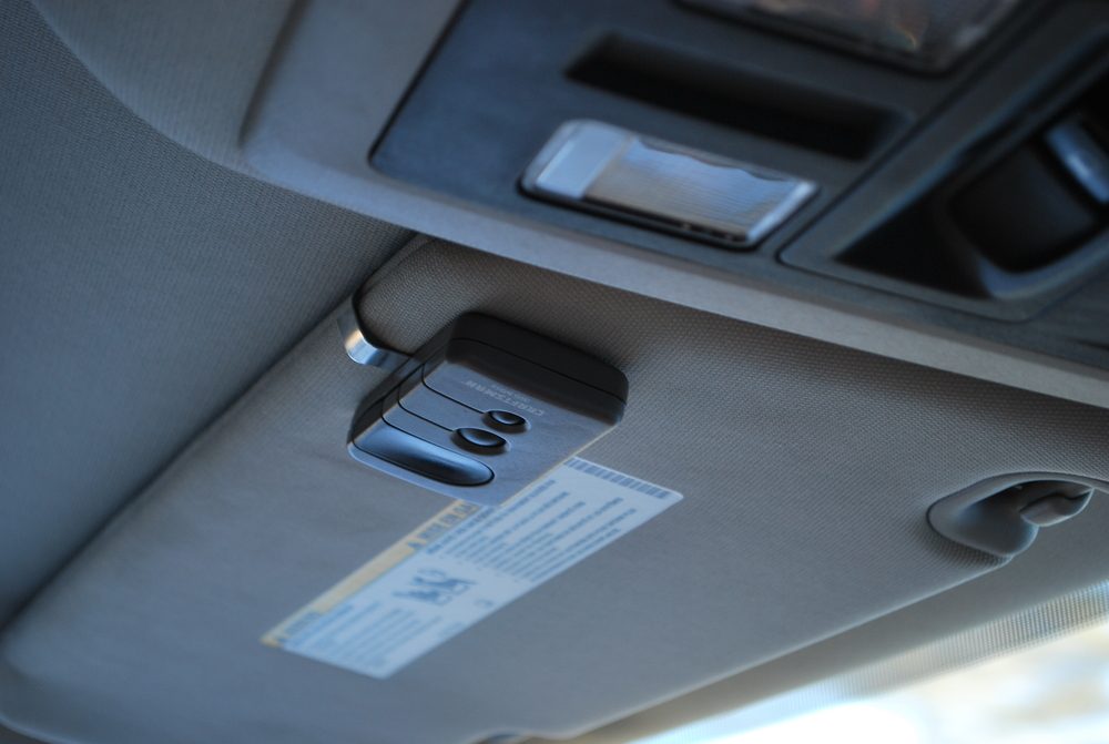 A car's sun visor displays a control panel with buttons and a warning label, mounted above the front windshield in a light-colored interior.