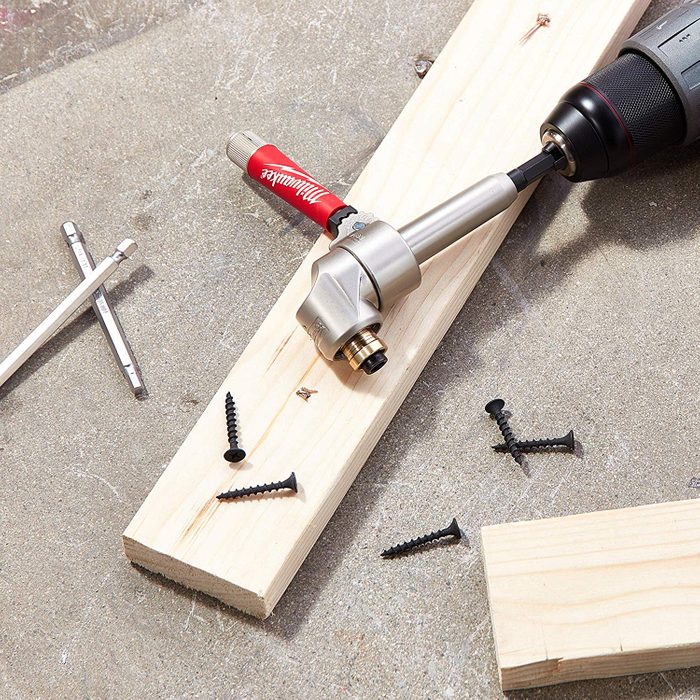 A drill attached to a drive adapter is positioned over a wooden plank, with black screws scattered nearby on a concrete surface.