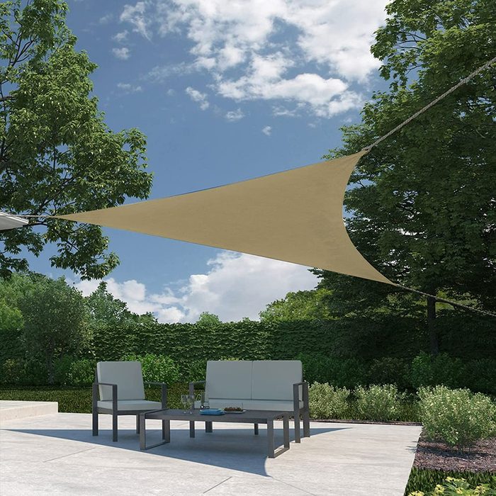 A triangular shade sails above a patio set, providing shelter. Green shrubs and trees surround the area under a partly cloudy blue sky.