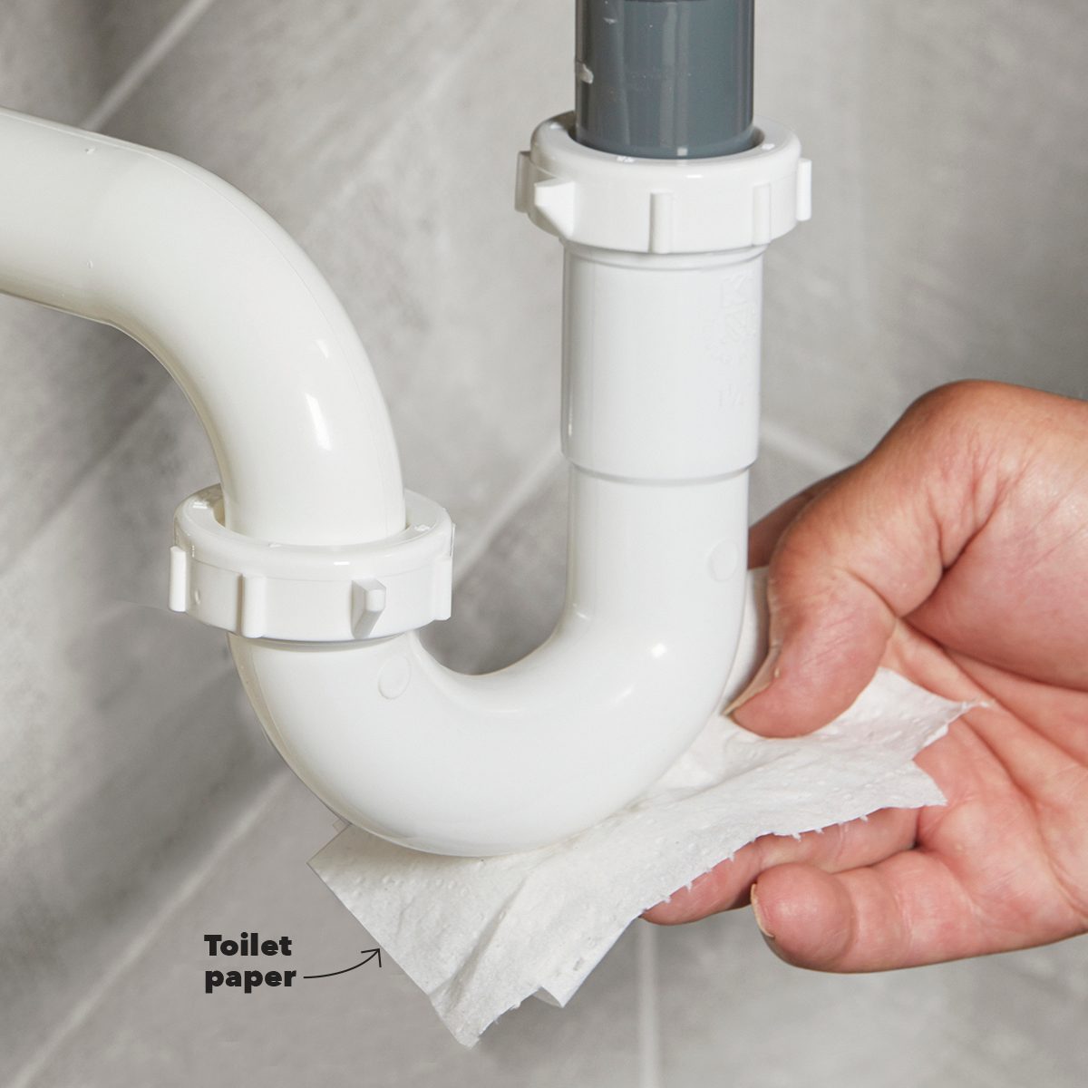 A hand holds a piece of toilet paper against a curved white pipe, likely checking for leaks, in a tiled bathroom setting.