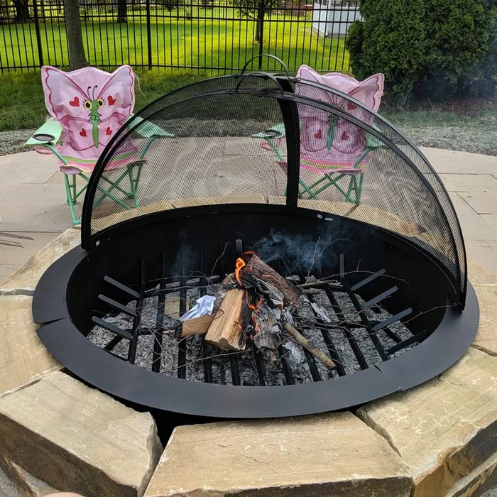 A fire burns in a circular metal pit, with smoke rising. Two butterfly-themed folding chairs sit nearby on a paved surface, surrounded by green grass.