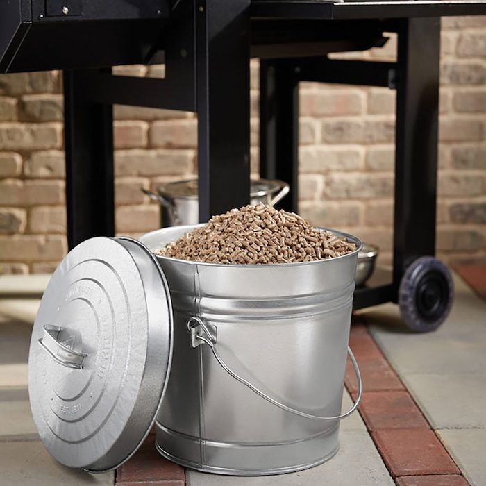 A silver bucket filled with wood pellets sits open next to a black grill, against a backdrop of a brick wall and paved ground.