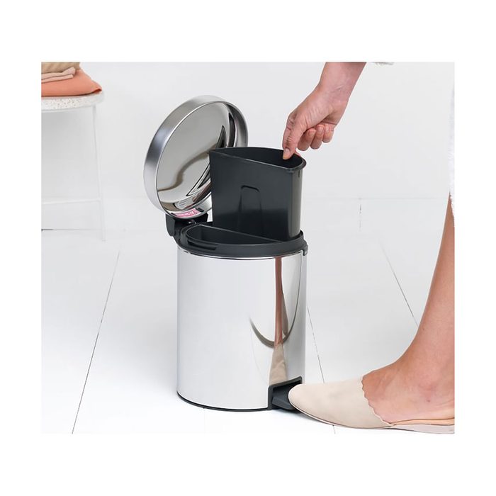A hand lifts a black inner bin from a shiny, foot-operated trash can in a bright, clean, minimalist room with a white floor.