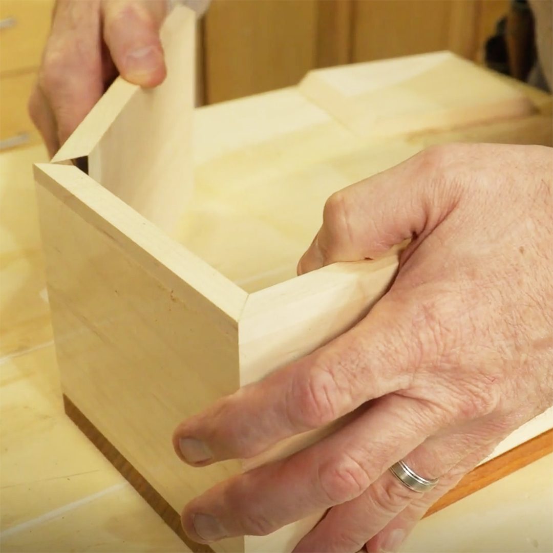 A person assembles wooden pieces together, gripping them with hands. The setting features a light-colored work surface and surrounding cabinetry.