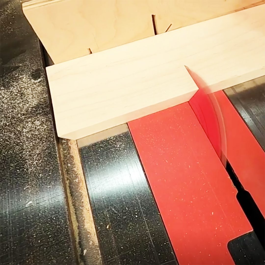 A wooden plank is being cut on a table saw with a red insert, surrounded by sawdust on a black surface.