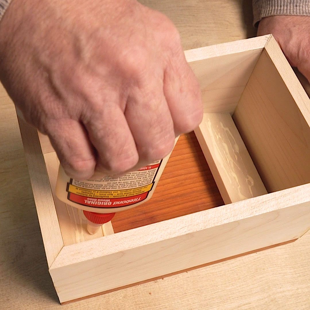 A hand squeezes glue into a wooden box, preparing to assemble its parts. The workspace features a light-colored wooden surface.
