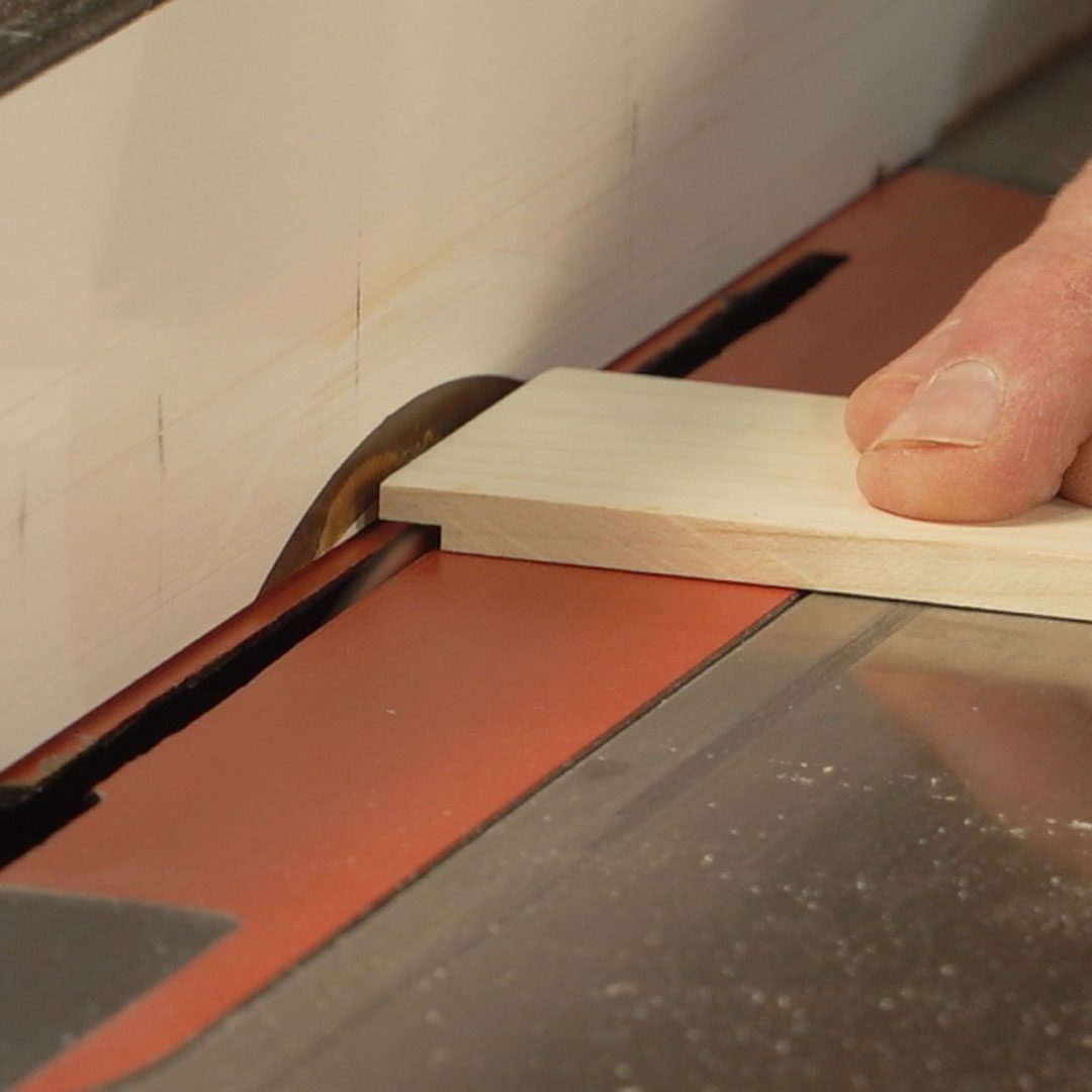 A hand holds a wood piece against a spinning saw blade, cutting it while positioned on a saw table in a workshop environment.